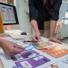 Two female ux designers are collaborating on a project, arranging wireframes on a table to design the user interface and experience for a mobile application